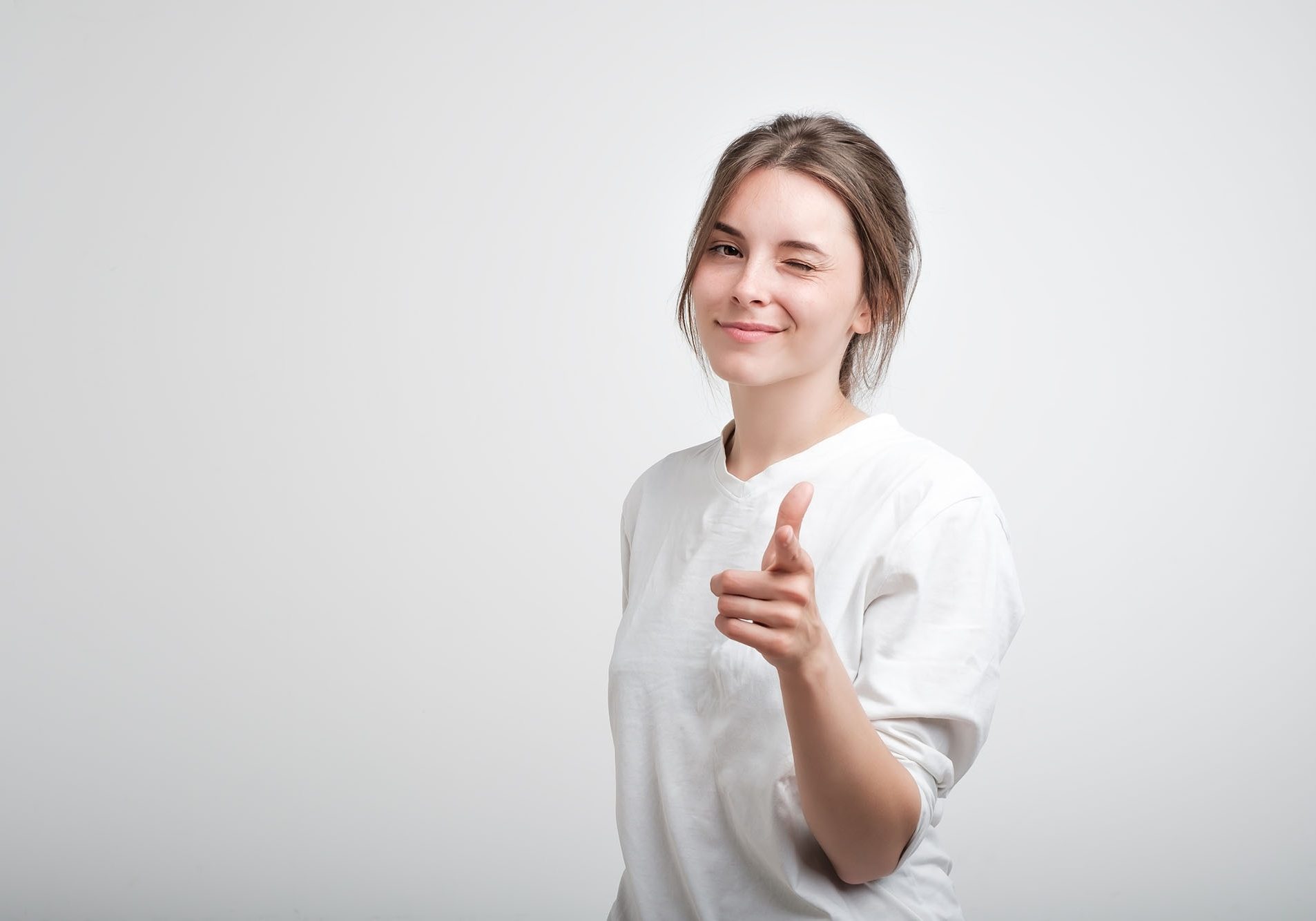 Positive cheerful young caucasian woman wearing white casual T-shirt blinking her eyes and smiling pointing at camera with index fingers. Facial emotion. Succees in life or studies